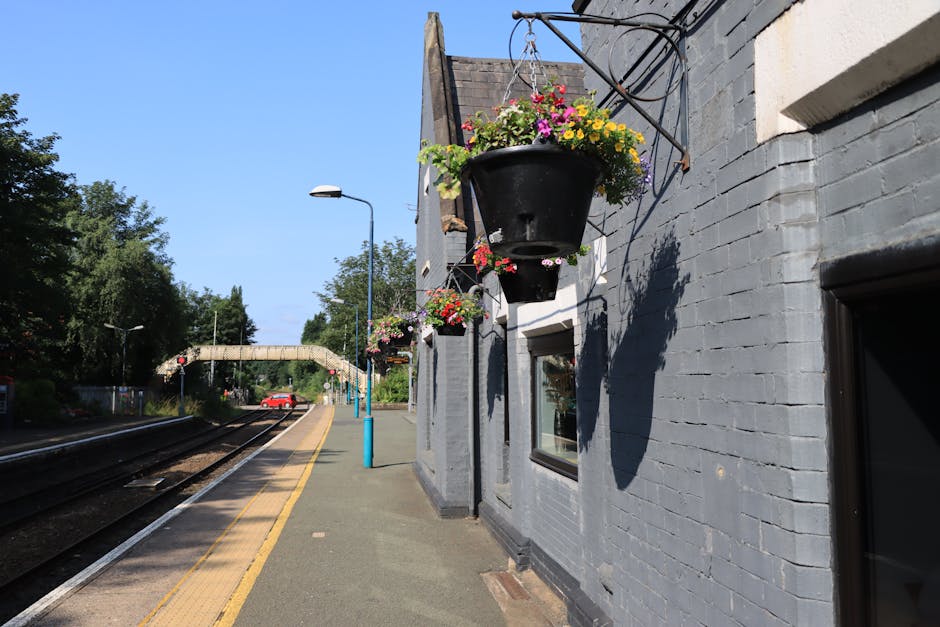 A close-up view of hanging flower baskets mounted on black iron brackets attached to a grey brick wall outside West Brompton station. The baskets contain vibrant, fresh flowers including pink, yellow,